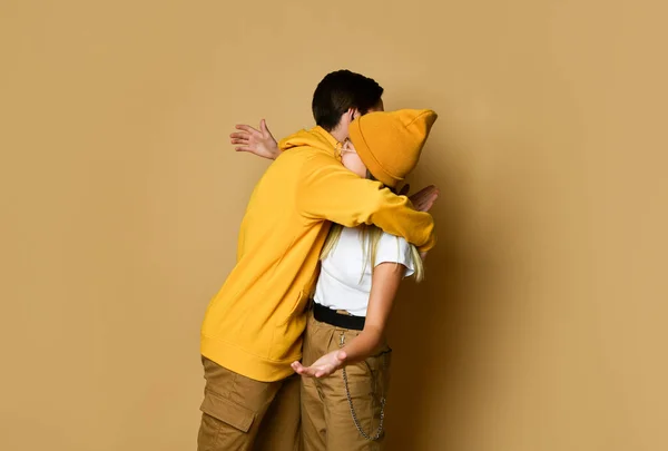 Young teens boy and girl in comfortable clothing, hats and sunglasses standing and embracing each other over yellow background — Stock Photo, Image