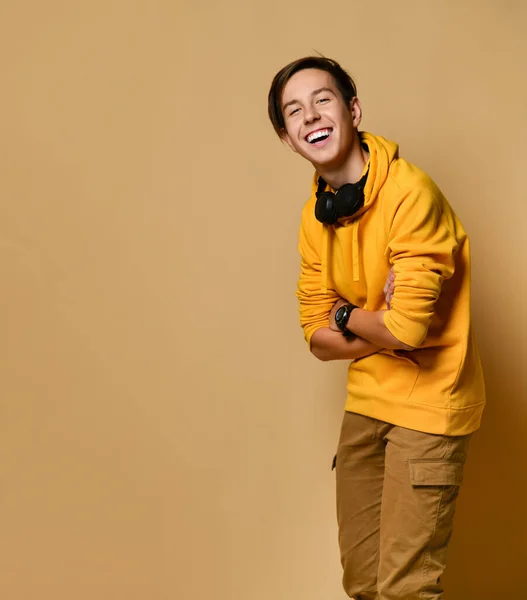 Joven adolescente positivo en sudadera con capucha amarilla y auriculares de pie, sonriendo y mirando a la cámara sobre fondo amarillo de la pared —  Fotos de Stock