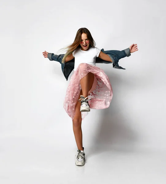 Young positive teen girl in stylish casual clothes and sneakers standing and moving with hands stretched out over white background — Stockfoto