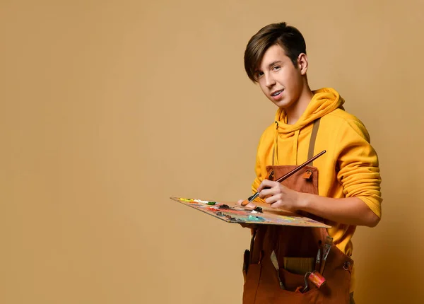 Jovem adolescente pintor de capuz amarelo e macacão de pé e pintura em cavalete sobre fundo amarelo — Fotografia de Stock