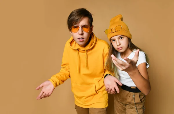 Adolescentes jóvenes niño y niña en ropa cómoda, sombreros y gafas de sol de pie y expresando malentendidos —  Fotos de Stock