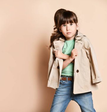 a little brunette girl in a beige raincoat, with her arms crossed over her chest