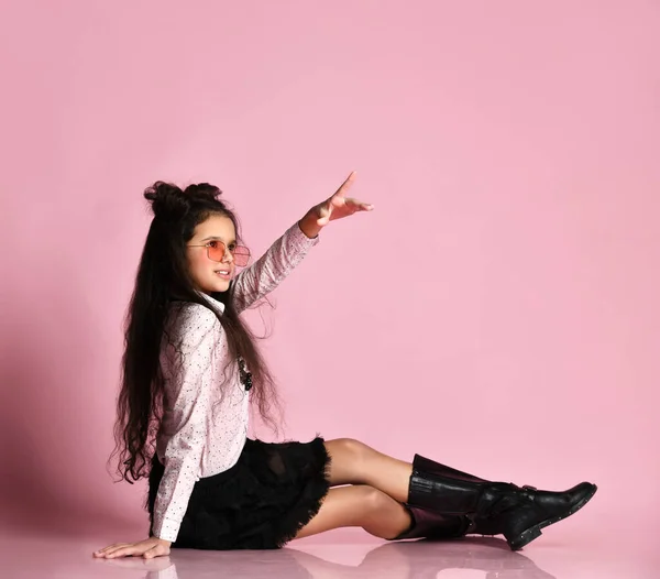 Tiener kind met lang haar, in een zonnebril, witte blouse, zwarte rok en laarzen. Poseren zittend op roze achtergrond. Sluiten. — Stockfoto