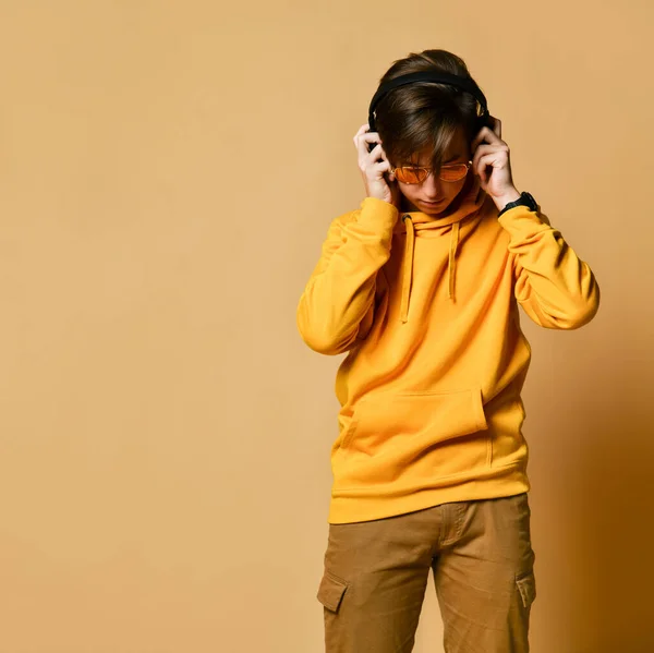 Jovem adolescente positivo com capuz amarelo, óculos de sol e fones de ouvido de pé e sorrindo — Fotografia de Stock