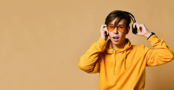Joven adolescente positivo en sudadera con capucha amarilla, gafas de sol y auriculares de pie y mirando a la cámara sobre fondo amarillo de la pared —  Fotos de Stock