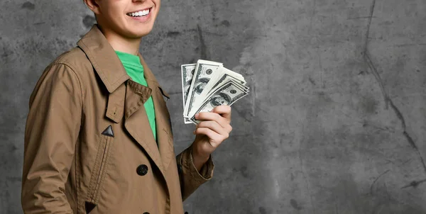 Young smiling teen boy in brown jacket and glasses standing and holding heap of dollars in hand over grey concrete wall background — ストック写真