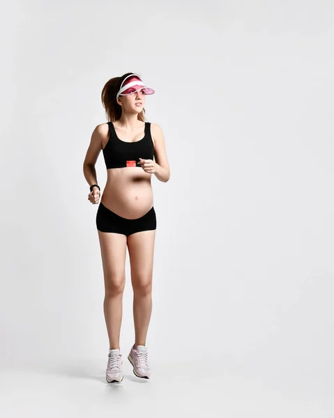 Yeşil güneşlikli hamile kız, siyah spor şortlu ve üstü pembe spor ayakkabılı. Beyazlar üzerinde tek başına koşuyor. Tam uzunluk — Stok fotoğraf