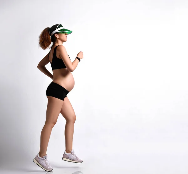 Fille enceinte en vert pare-soleil, short de sport noir et haut, baskets roses. Elle fait du jogging isolée sur du blanc. Longueur totale — Photo