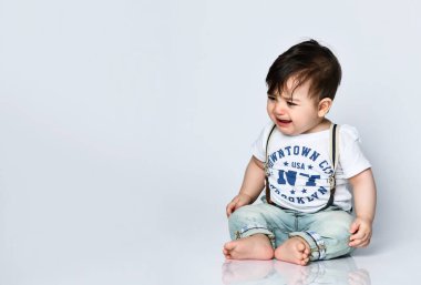 Üzerinde yazılar olan tişörtlü bir çocuk pantolon pantolon pantolon ve patik. Yerde oturuyor ve ağlıyor, beyaz bir arka planda tek başına.