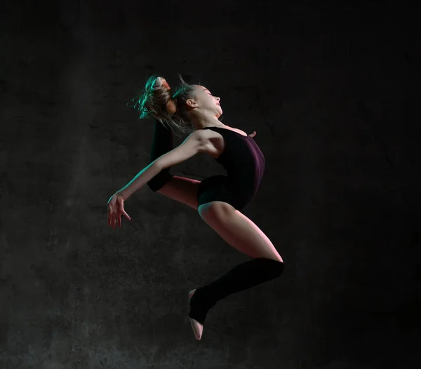 Chica joven gimnasta en cuerpo deportivo negro y la parte superior especial saltando en pose gimnástica sobre fondo oscuro — Foto de Stock