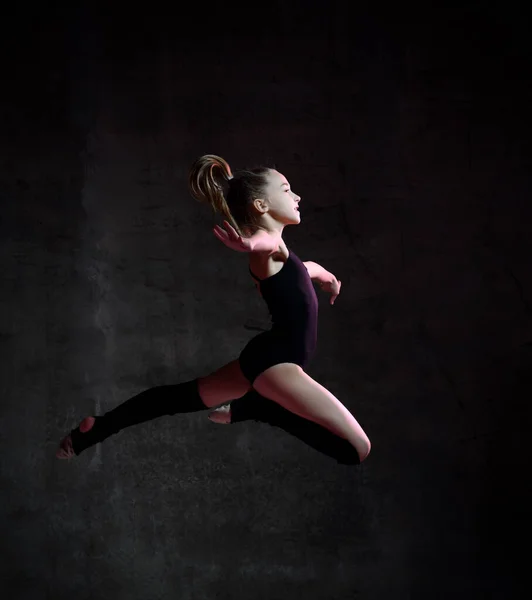 Junge Turnerin in schwarzem Sportbody und speziellen Oberteilen, die in Turnpose vor dunklem Hintergrund springen — Stockfoto