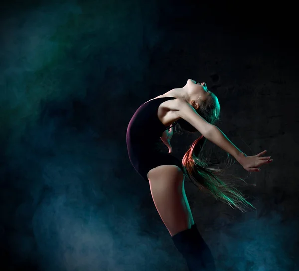 Young girl gymnast in black sport body and special uppers jumping in gymnastic pose over dark background — Stock Photo, Image
