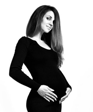 Dar elbiseli hamile bir kadının siyah beyaz fotoğrafı. Ellerini karnına koydu, beyaz arka planda izole bir şekilde poz verdi. Kapat.