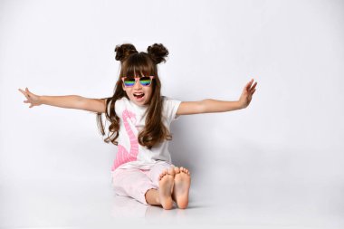 Stüdyoda, güneş gözlüklü ve şık saç stili olan çıplak ayaklı bir kız poz veriyor.