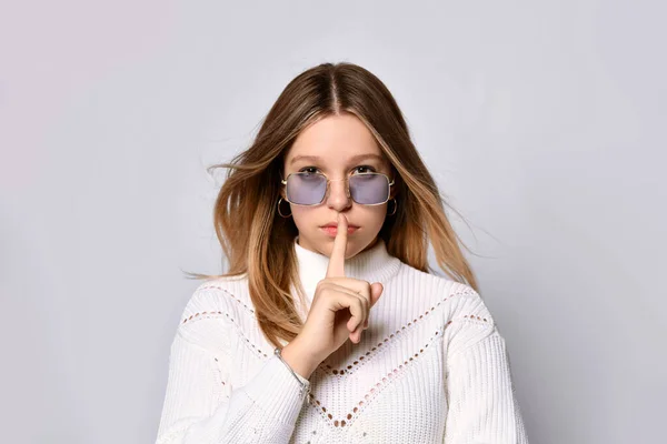 Blonde adolescent in sunglasses, smart watch, bracelet and sweater. She showing be quiet sign, posing isolated on white. Close up — Stock Photo, Image