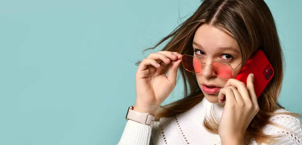 Adolescente en gafas de sol naranjas, reloj, pulsera y suéter. Hablaba por teléfono, sorprendida, posando sobre fondo azul. De cerca. — Foto de Stock