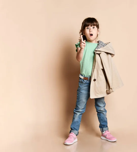 Roztomilá dívka pózující v různých pózách, v džínách s opaskem, zeleným tričkem a béžovým kabátem a růžovými tenisky — Stock fotografie