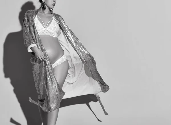 Bianco nero foto ritagliata di una donna incinta sexy in lingerie sexy e un accappatoio scintillante d'oro. Cammina su uno sfondo beige. Famiglia e maternità, moda e bellezza — Foto Stock