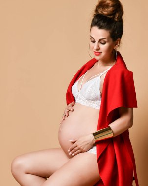 Küpeli esmer anne, bilezik, beyaz iç çamaşırı ve kırmızı şal. Sandalyeye yan oturup, bej arka planda poz vermek. Yakın plan.