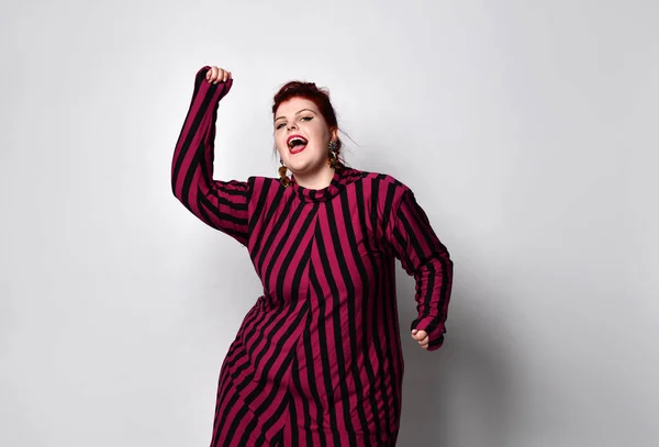 Overgewicht roodharige vrouw in gestreepte jurk, oorbellen. Ze danst en zingt terwijl ze poseert op een witte foto achtergrond — Stockfoto