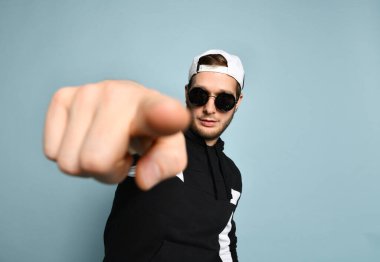Hipster male in cap, black tracksuit and sunglasses. He is pointing at you by his forefinger, posing on blue background. Close up