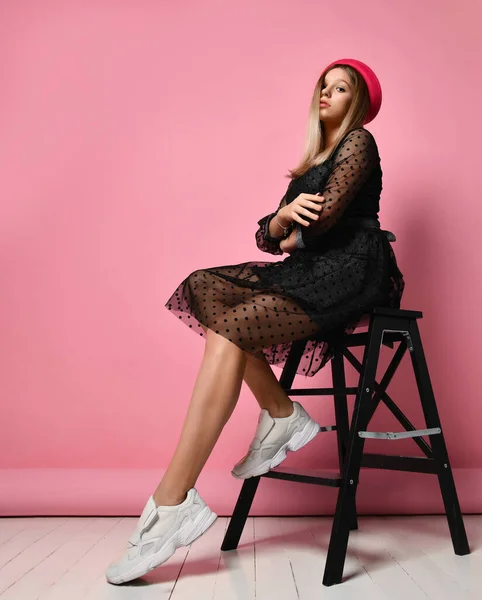 Menina adolescente em vestido preto, chapéu vermelho e tênis brancos. Ela sentou-se na escada do degrau, posando de lado no fundo rosa. Comprimento total — Fotografia de Stock