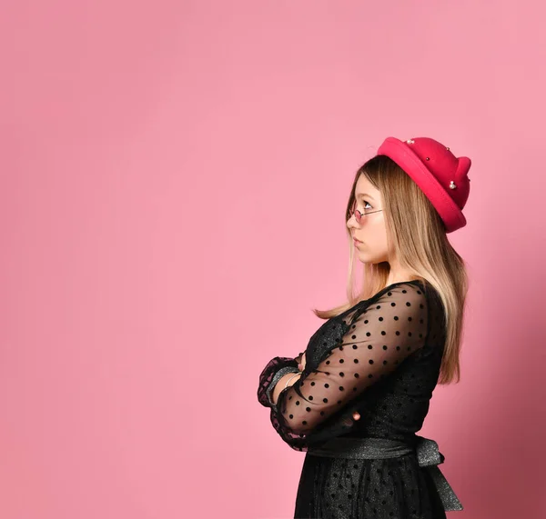 Adolescente em óculos de sol, vestido preto e chapéu vermelho. Ela está olhando para cima, mãos dobradas, posando de lado no fundo rosa. Fechar — Fotografia de Stock