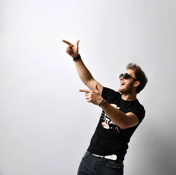 Hipster companheiro de óculos de sol, t-shirt preta com estampa, pulseiras. Ele está apontando para alguém, posando isolado no branco. Fechar — Fotografia de Stock