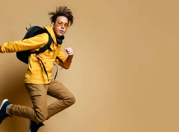 Νεαρό έφηβος αγόρι σε άνετα ρούχα, πάνινα παπούτσια, γυαλιά ηλίου και σακίδιο άλμα πάνω από κίτρινο φόντο τοίχο — Φωτογραφία Αρχείου