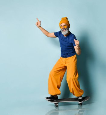 Tişörtlü, güneş gözlüklü, turuncu pantolonlu, şapkalı, spor ayakkabılı yaşlı bir adam. Siyah kaykay sürmek, yumruklarını göstermek, mavi arka planda poz vermek.