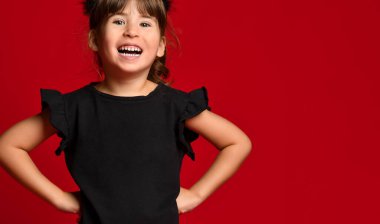 Kırmızı arka planda siyah tişörtlü tatlı esmer kız. Bebek ellerini kalçalarına koydu ve kameraya gülümsedi..