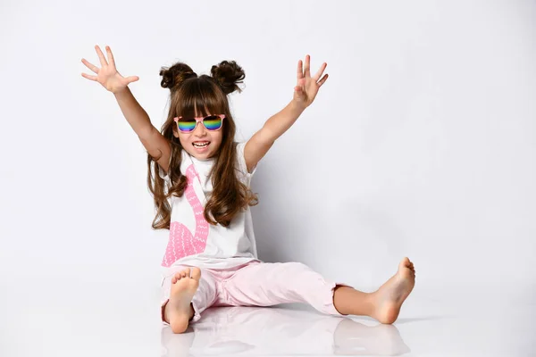 Маленька весела дівчинка позує сидячи на підлозі, в студії, в темних окулярах і модних зачісках, босоніж — стокове фото