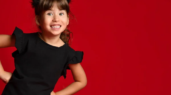 Schattig brunette meisje in een zwart T-shirt op een rode achtergrond. De baby legde haar handen op haar heupen en lacht naar de camera. — Stockfoto
