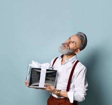 Beyaz gömlekli, kahverengi pantolonlu ve pantolon askılı gri saçlı adam. Gülümseyen, elinde gümüş bir hediye kutusu olan, mavi arka planda poz veren