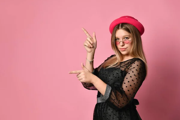 Adolescente rubia en gafas de sol, vestido negro y sombrero rojo. Ella sonrió, señalando algo, posando sobre un fondo rosa. De cerca. — Foto de Stock