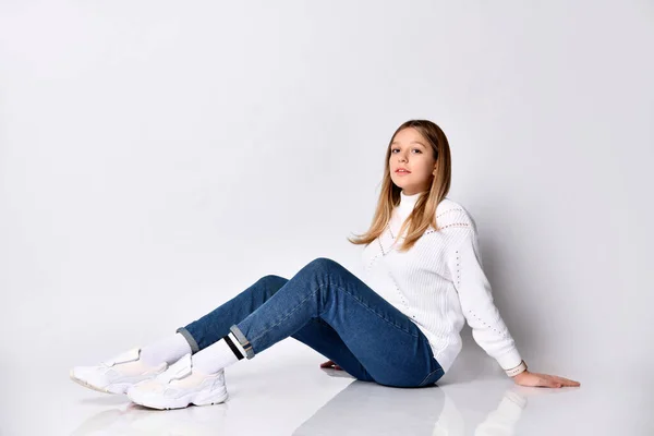 Adolescente com cabelo comprido. Vestida com jeans, camisola, meias e tênis. Está sentada no chão, isolada em branco. Fechar — Fotografia de Stock