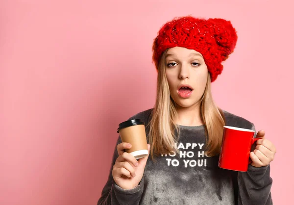 Ragazza in pigiama grigio renna, cappello rosso. Ha in mano tazze di ceramica e di carta, cercando assonnato, in posa su sfondo rosa. Da vicino. — Foto Stock