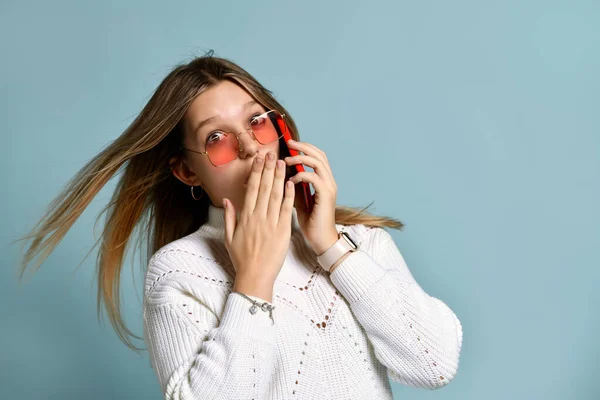 Adolescent in orange sunglasses, watch, bracelet and sweater. She talking by phone, surprised, posing on blue background. Close up — Stock Photo, Image