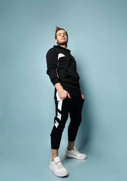 Hipster male in black tracksuit, bracelet and sneakers. He is rolling up his sleeve, posing against blue background. Full length