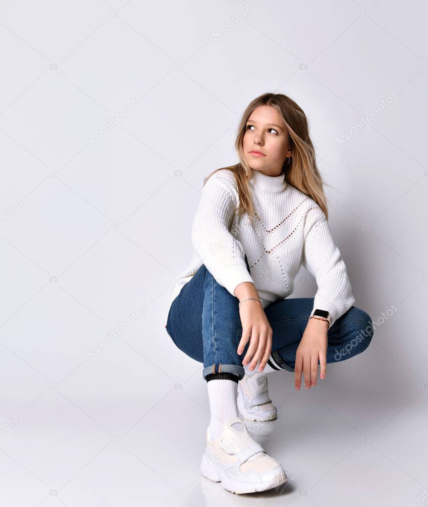 Teenage girl with long hair. Dressed in jeans, sweater and sneakers. Kneeling on floor and smiling, isolated on white. Close up