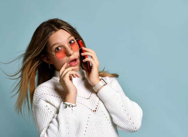 Adolescente in occhiali da sole arancioni, orologio, bracciale e maglione. Lei parla per telefono, sorpreso, in posa su sfondo blu. Da vicino. — Foto Stock