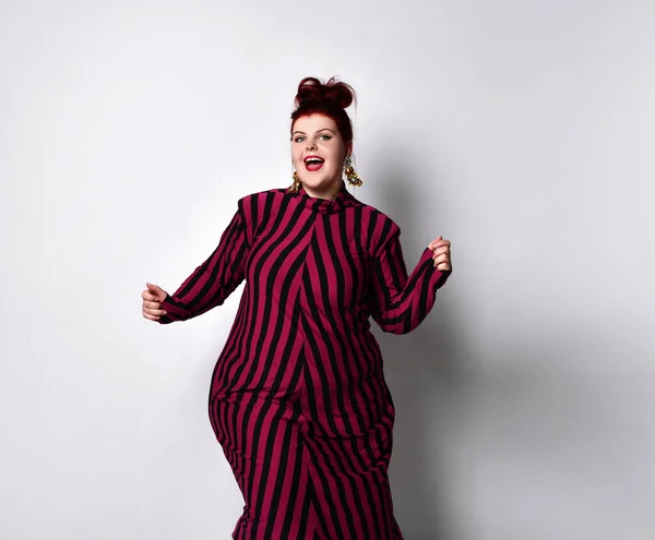 Obese redhead lady in striped dress and earrings. She reaching her hand to you while posing sideways isolated on white background — Stockfoto