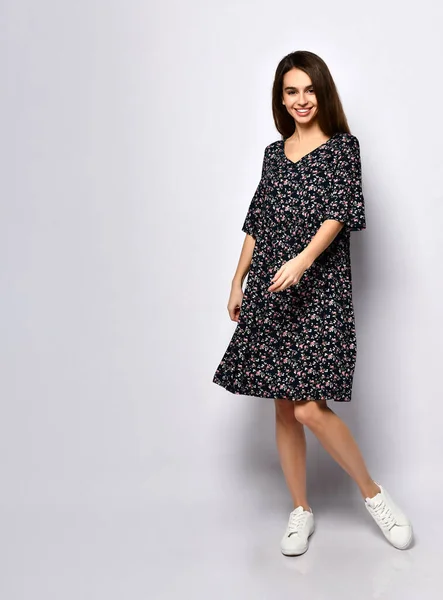 Preciosa mujer morena en vestido negro con estampado floral y zapatillas blancas. Sonriendo, posando aislada sobre blanco. Longitud completa —  Fotos de Stock