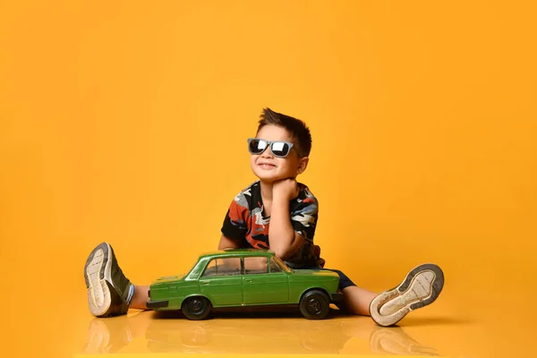 Dzieciak w okularach przeciwsłonecznych, koszulce z kamuflażem, tenisówkach. Siedzenie na podłodze, trzymanie zielonego modelu samochodu retro, pozowanie na pomarańczowym tle — Zdjęcie stockowe