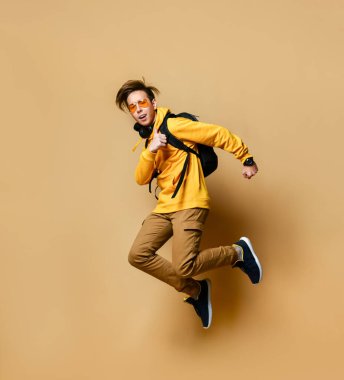 Young teen boy in comfortable clothing, sneakers and backpack jumping over yellow wall background