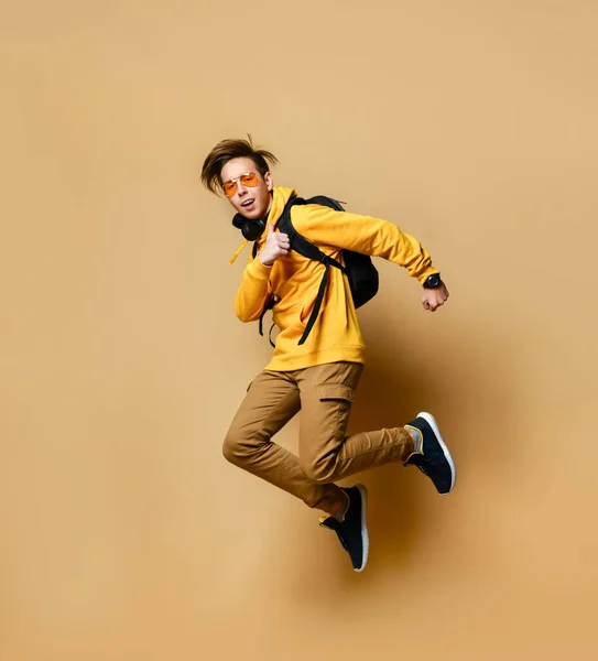 Young teen boy in comfortable clothing, sneakers and backpack jumping over yellow wall background