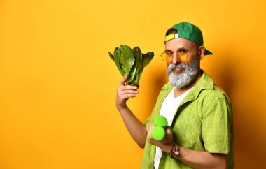 Yeşil şapkalı, gömlekli, beyaz tişörtlü, güneş gözlüklü yaşlı bir adam. Turuncu arka planda iki dambıl ve marul duruyor.