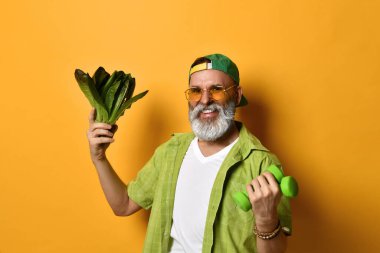 Yeşil şapkalı, gömlekli, beyaz tişörtlü, güneş gözlüklü yaşlı bir adam. Turuncu arka planda iki dambıl ve marul duruyor.