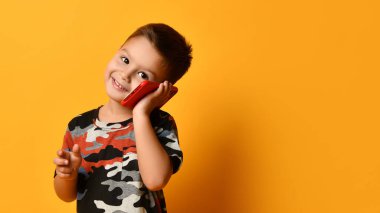 Kamuflaj tişörtlü küçük esmer çocuk gülümsüyor, kırmızı akıllı telefonla konuşuyor, sana bakıyor, turuncu arka planda poz veriyor.
