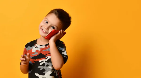 stock image Little brunet boy in camouflage t-shirt is smiling, talking by red smartphone, looking at you, posing against orange background